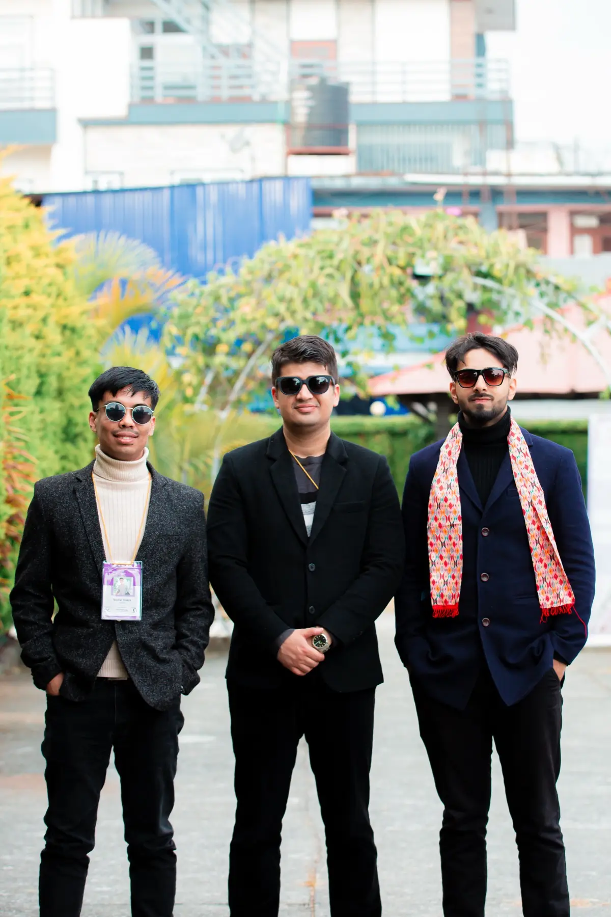 Three young men in dark jackets with event lanyards, one wearing a patterned scarf, standing together outdoors