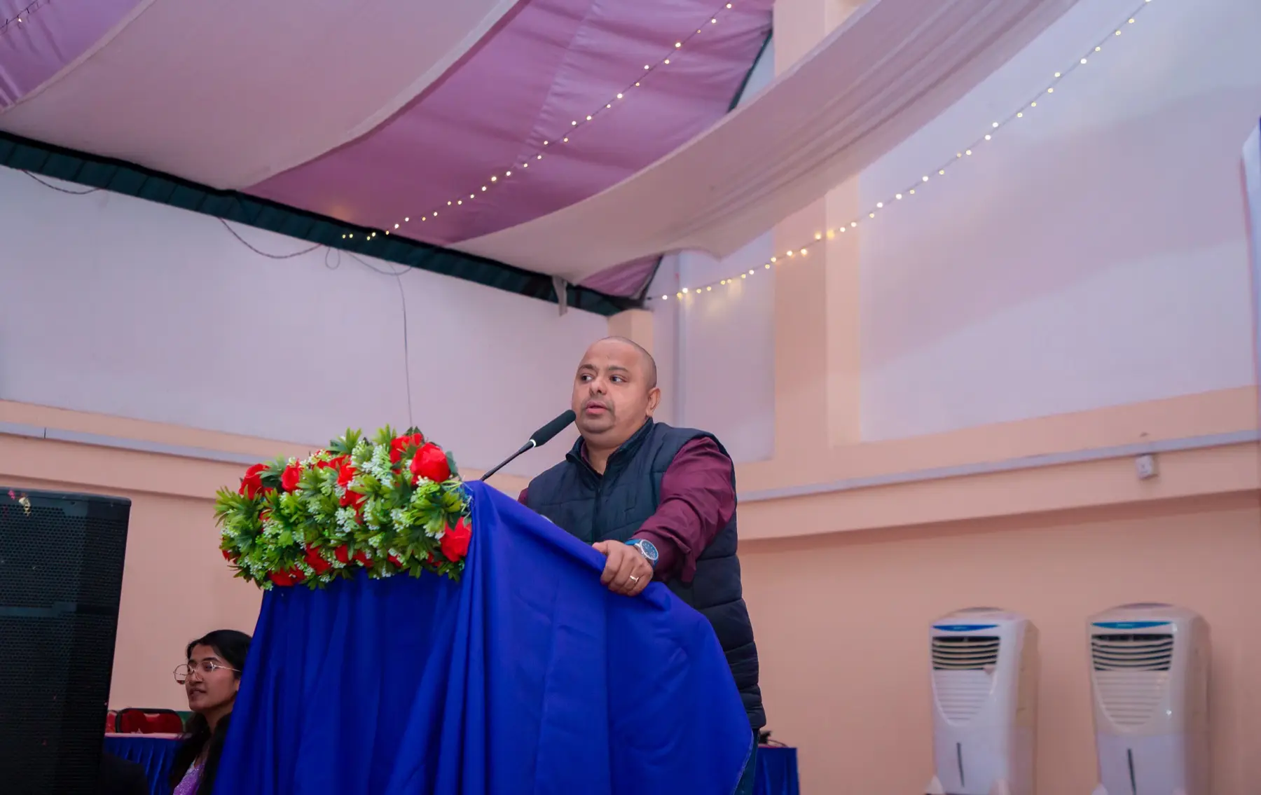Speaker leaning on a blue-draped podium with flowers, with a listener visible at the lower left