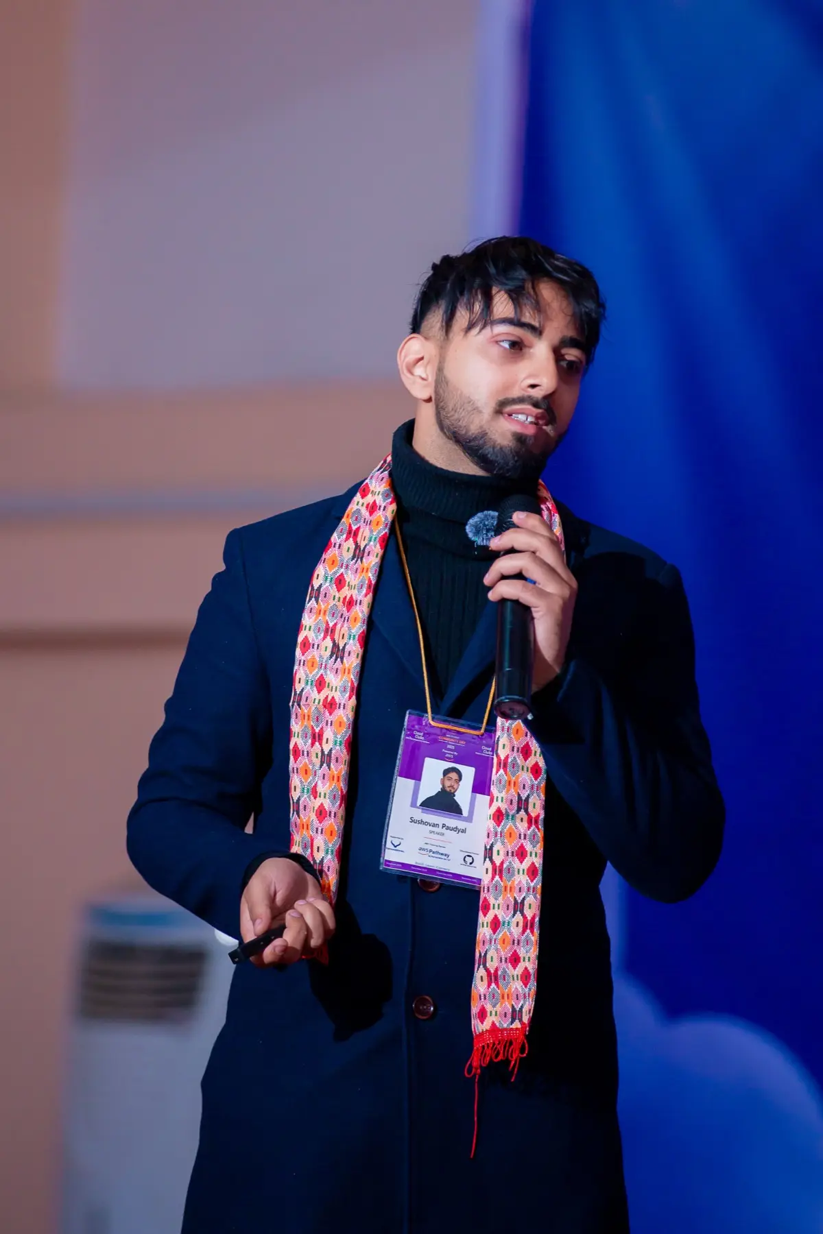 Speaker with a patterned scarf and event badge talking into a handheld microphone on stage