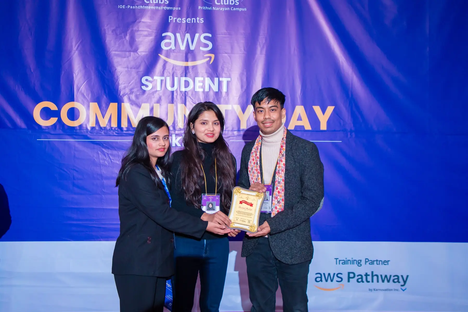 Three people on stage holding a golden plaque together in front of the AWS Student Community Day backdrop