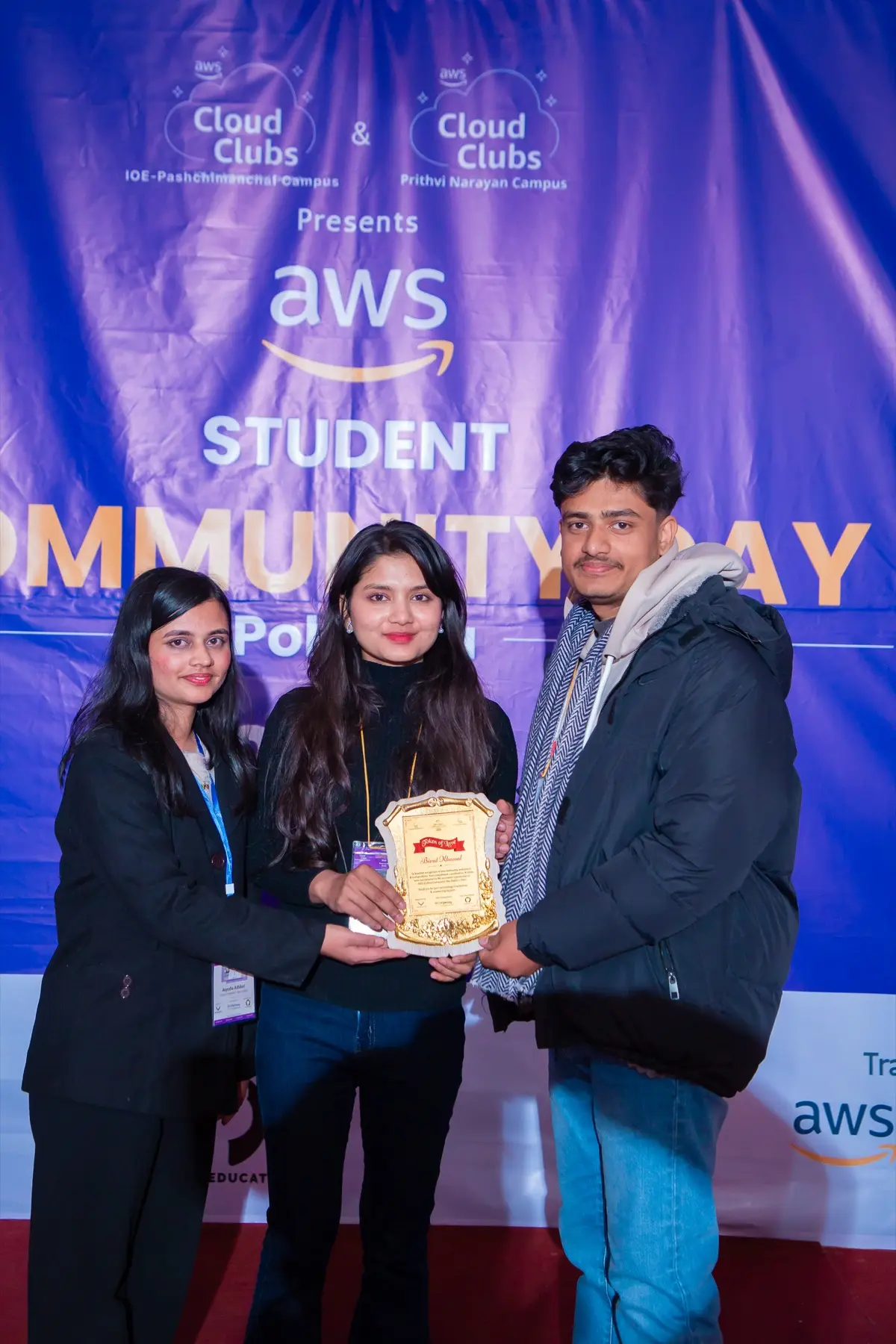 Three people receiving a golden plaque in front of the AWS Student Community Day backdrop