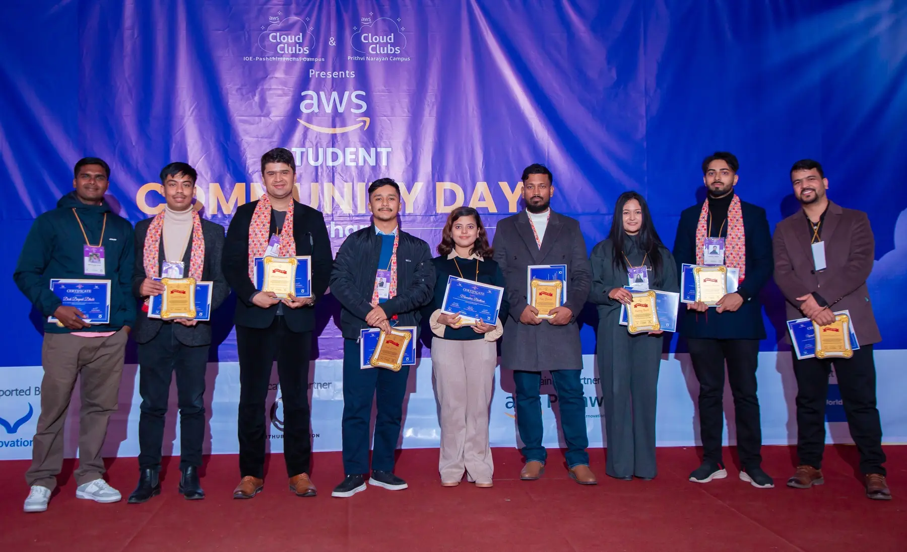Row of eight people standing on a red carpet holding certificates and plaques in front of the event backdrop